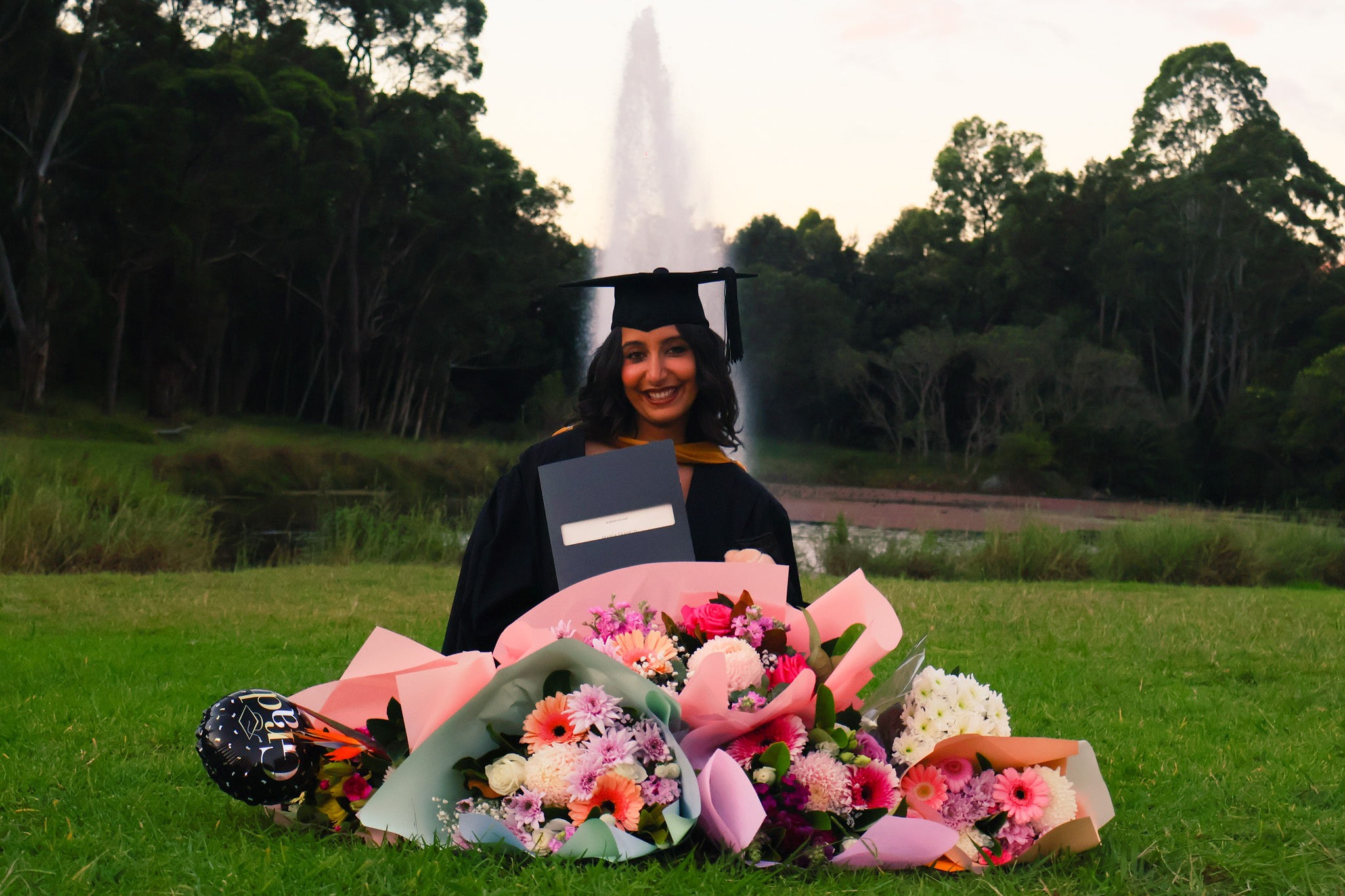 Graduate sitting behind bouquets of flowers near a fountain